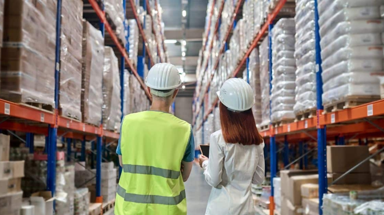 Two workers in hard hats and safety gear stand side by side in a large warehouse, looking up at tall shelves filled with stacked pallets of goods. One worker is wearing a neon yellow safety vest, while the other is dressed in a white shirt, holding a tablet.