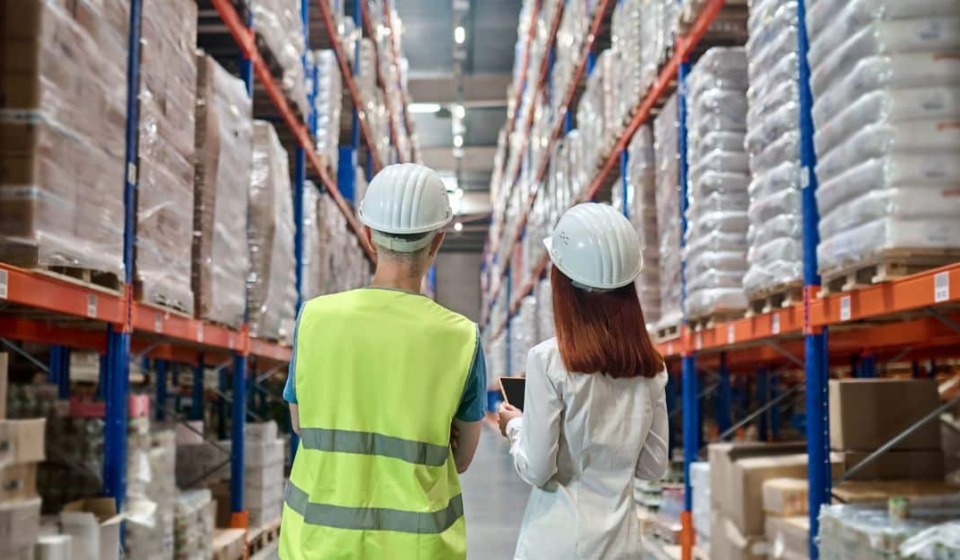 Two workers in hard hats and safety gear stand side by side in a large warehouse, looking up at tall shelves filled with stacked pallets of goods. One worker is wearing a neon yellow safety vest, while the other is dressed in a white shirt, holding a tablet.