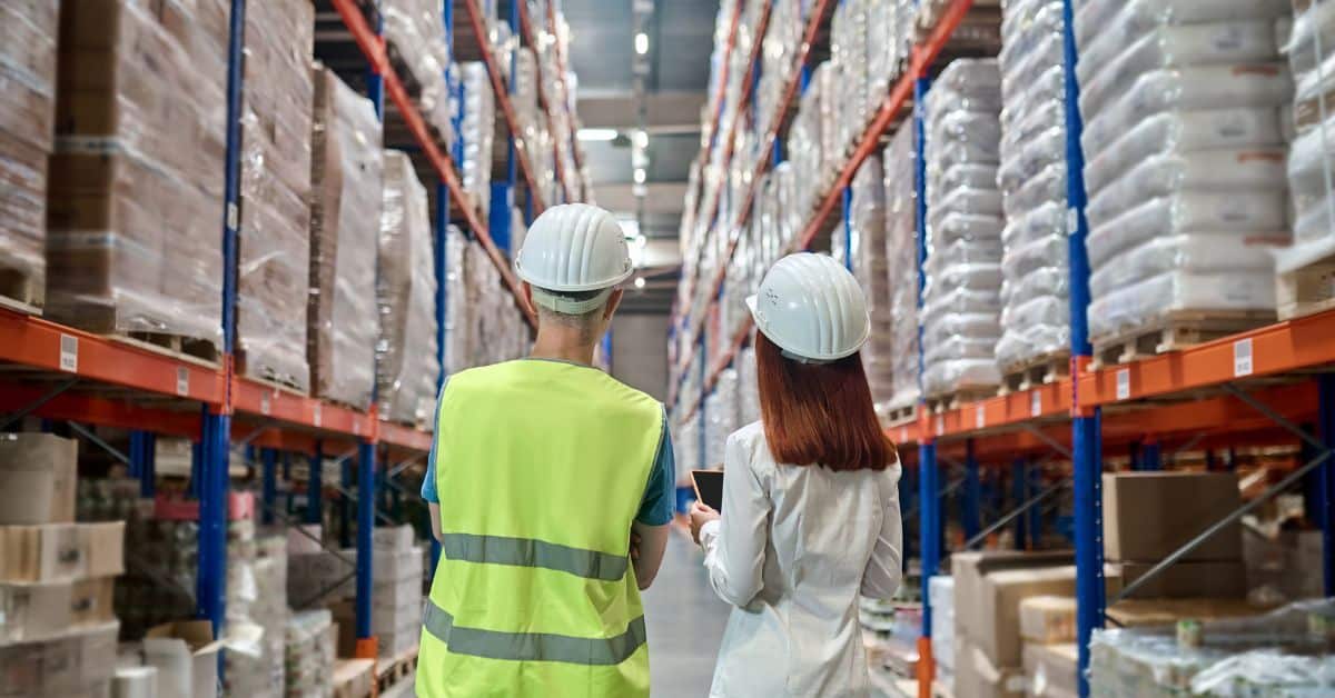 Two workers in hard hats and safety gear stand side by side in a large warehouse, looking up at tall shelves filled with stacked pallets of goods. One worker is wearing a neon yellow safety vest, while the other is dressed in a white shirt, holding a tablet.